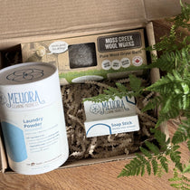 Meliora's laundry powder, a box with 3 wool dryer balls and a stain remover stick in a box filled with shredded brown paper and green leaves from a plant sitting on the right side of the box.