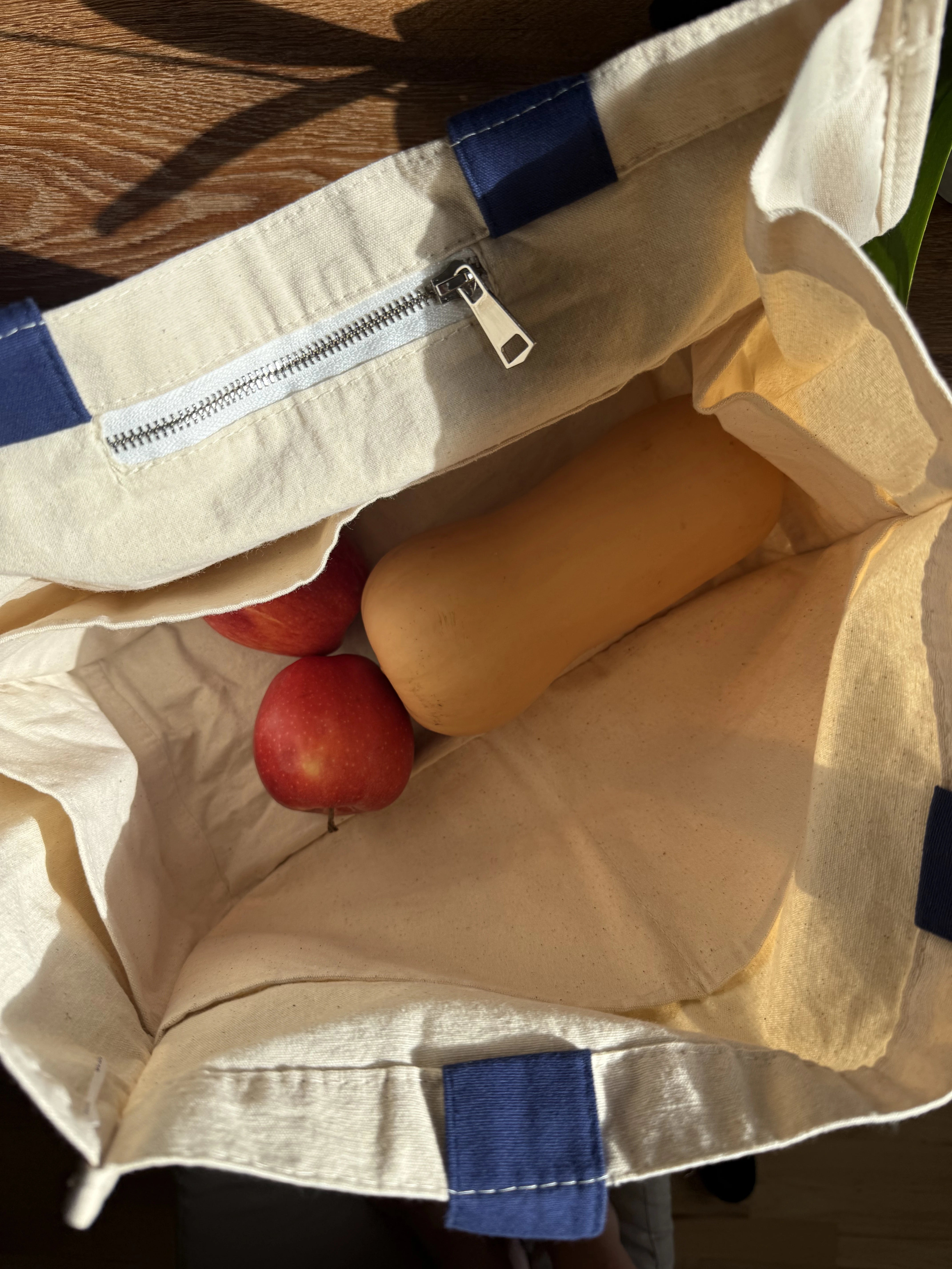 Beige reusable tote bag with blue handles containing multiple pockets and a pocket with a zipper, filled with 2 apples and a butternut squash.