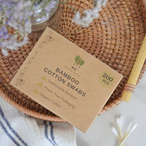 A kraft paper box of bamboo cotton swabs is displayed on a round woven wicker tray, surrounded by decorative props including dried lavender, a striped cloth, and cotton swabs.