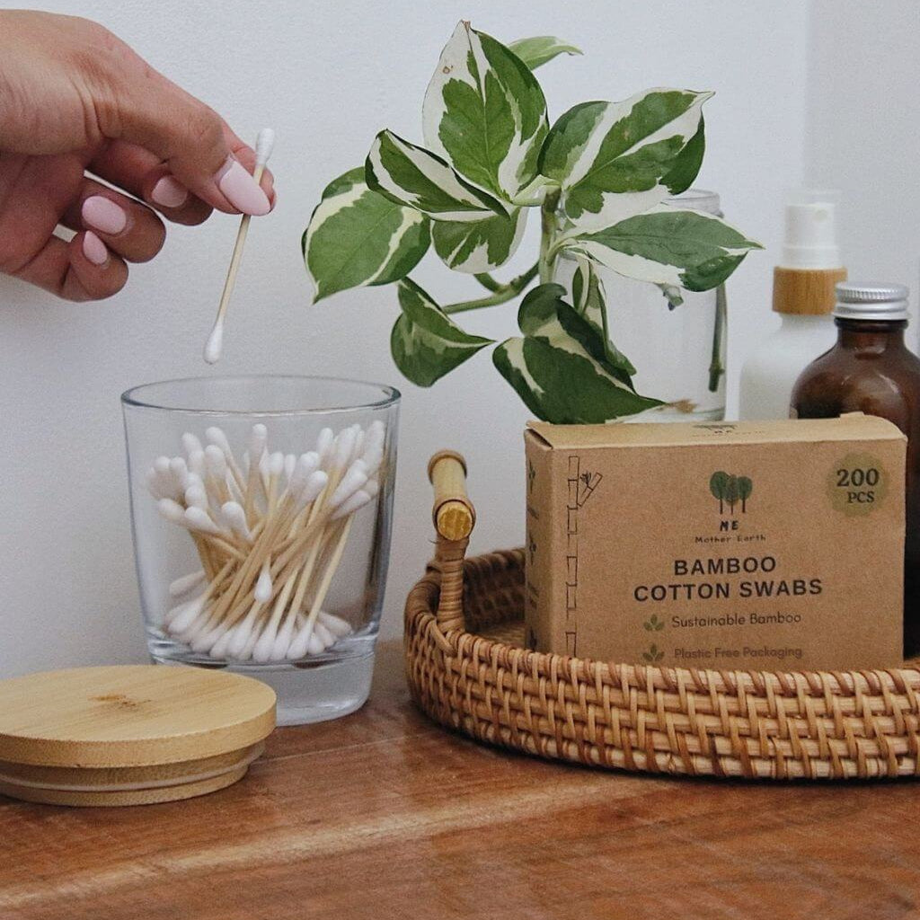 A hand with pink manicured nails holds a cotton swab above a glass jar filled with cotton swabs on a wooden table. A kraft paper box labeled 'BAMBOO COTTON SWABS' sits in a woven tray beside a green variegated plant and small bottles in the background.