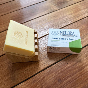 A bar of pale yellow Meljora Organic Products bath and body soap resting on a wooden stand next to its white and green cardboard packaging box, placed on a wooden deck floor.
