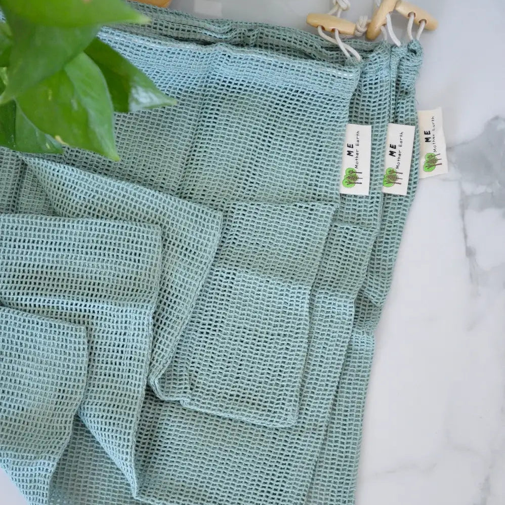 A stack of sage green waffle-weave reusable produce bags with drawstring closures, displayed on a white marble surface with green leaves in the foreground.