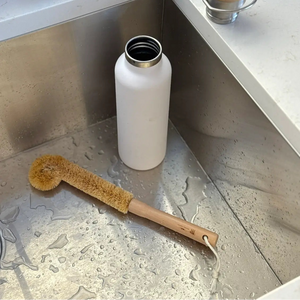 A matte white reusable bottle with a silver rim sits in a stainless steel kitchen sink, with a natural bristle brush and soap accessories nearby on a light countertop.