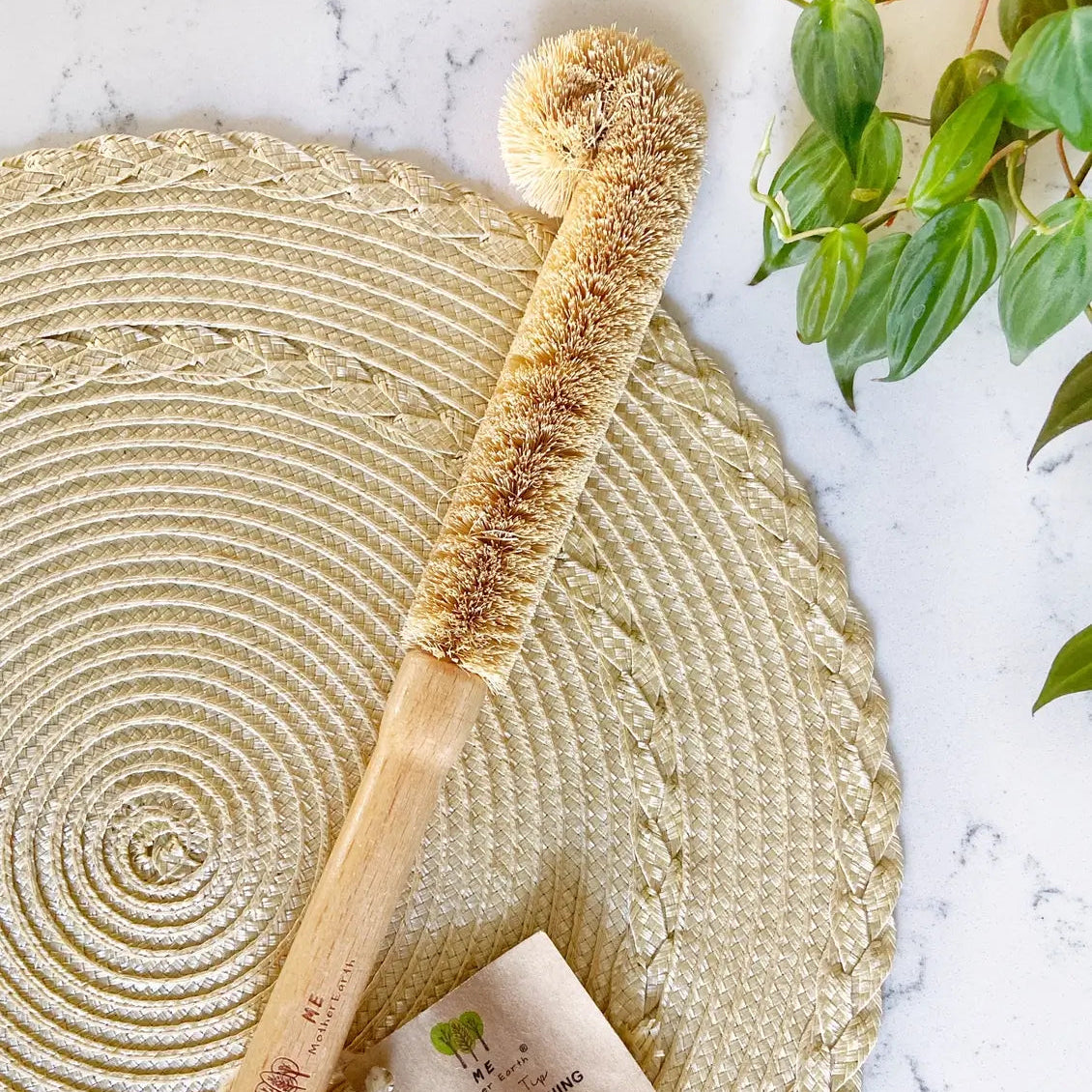 A natural coconut shell bottle cleaning brush with a bamboo handle and a small paper tag, styled on a round woven placemat over a white marble surface with green leaves in the corner.