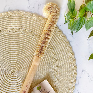 A natural coconut shell bottle cleaning brush with a bamboo handle and a small paper tag, styled on a round woven placemat over a white marble surface with green leaves in the corner.