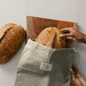 Person taking a loaf of bread out of a reusable bag with a wooden cutting board and another loaf of bread in the background.