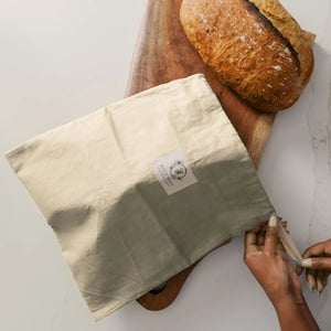 Person opening a beige bread bag with a loaf of bread on a wooden board.