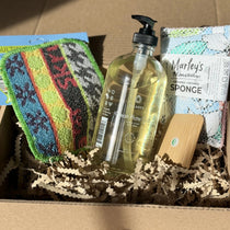 A set including a scrubbing pad, a washable sponge, a bamboo pot scrapper and a glass bottle of dish soap in a box filled with shredded brown paper