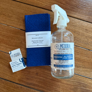 Melura Glass Cleaner kit on a wooden floor, including a clear spray bottle, a blue microfiber cloth, two absorbent tablets, and two test strips.