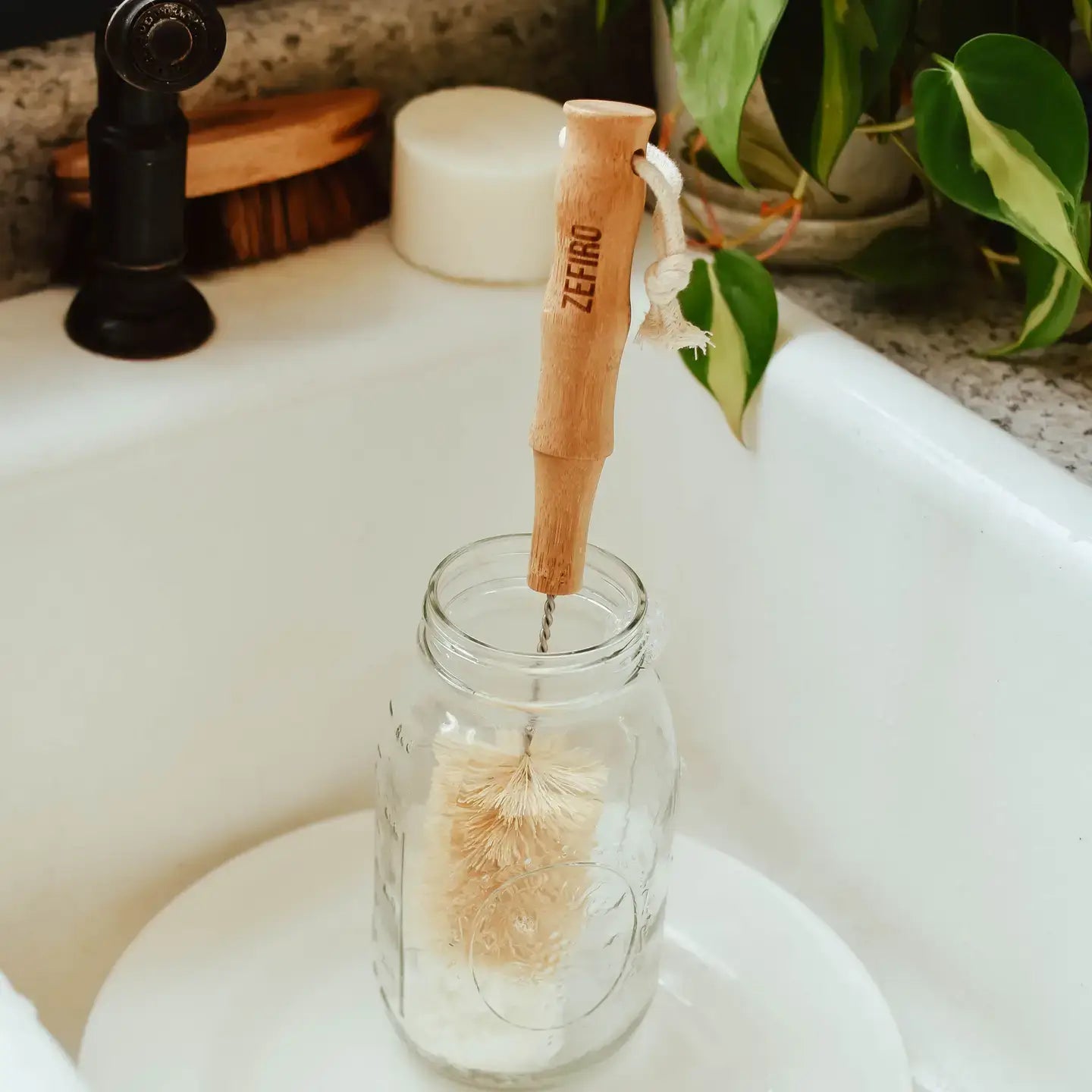 Bamboo dish brush in a glass jar on a sink with a plant in the background