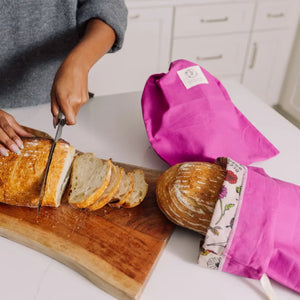 Person slicing bread on a wooden cutting board with pink oven mitts nearby.