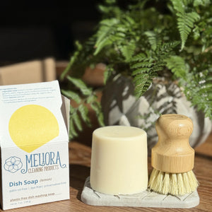 Meliora dish soap bar and its open packaging, bamboo dish brush brush, on soap dish on a wooden surface with plants in the background