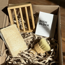 A solid dish block with a bamboo soap dish, a bamboo dish brush and a biodegradable sponge made with loofah and cellulose, all in a box filled with shredded brown paper.