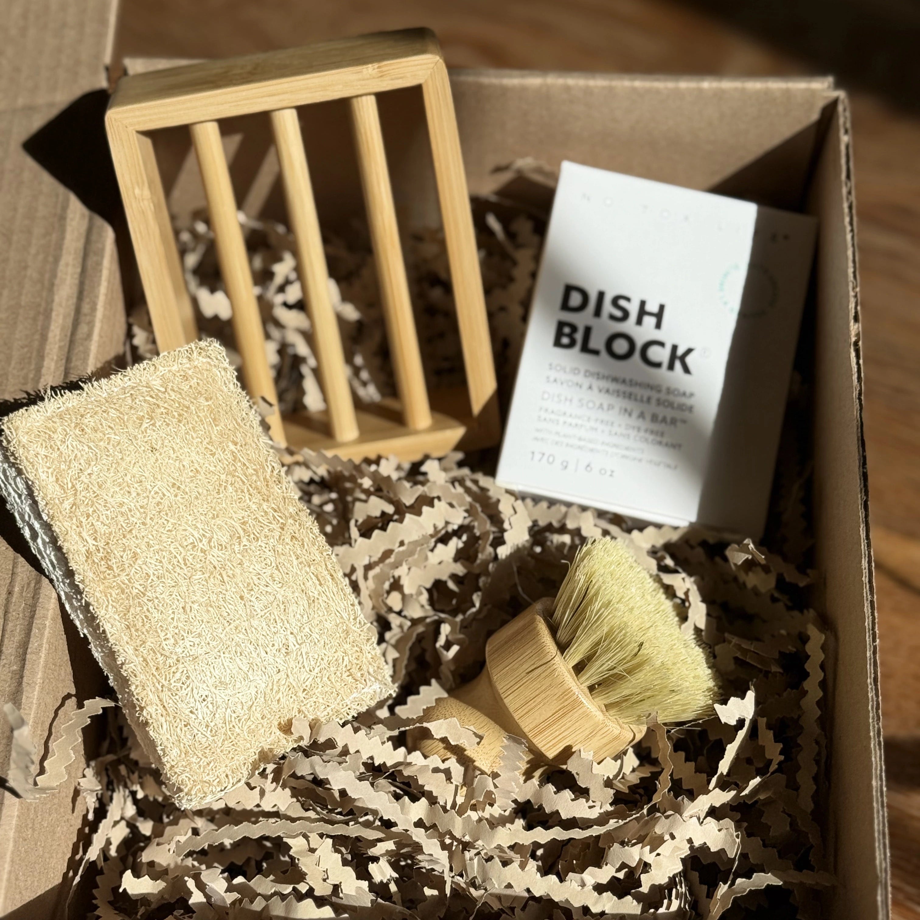 A solid dish block with a bamboo soap dish, a bamboo dish brush and a biodegradable sponge made with loofah and cellulose, all in a box filled with shredded brown paper.