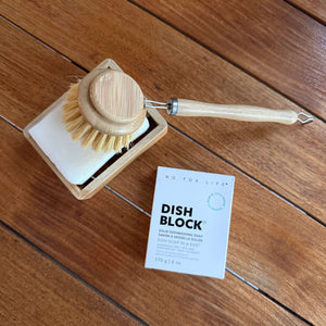 A wooden-handled dish brush with natural bristles resting on a square dish cleaning block, alongside a small white product card labeled 'DISH BLOCK' on a wooden floor.