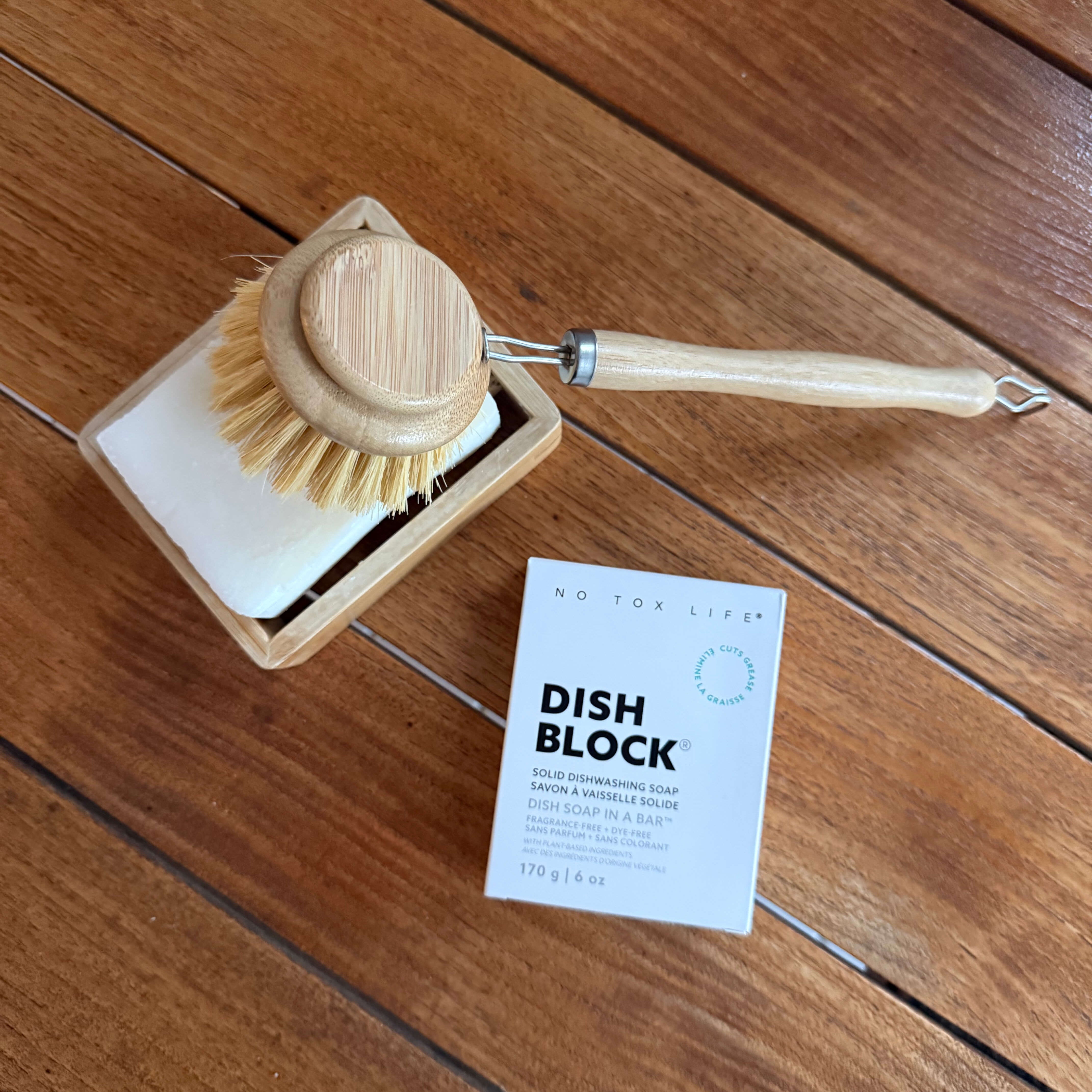 A wooden-handled dish brush with natural bristles resting on a square dish cleaning block, alongside a small white product card labeled 'DISH BLOCK' on a wooden floor.