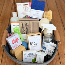 A curated bath and body gift basket arranged in a gray fabric basket on a wooden deck. The basket contains a variety of personal care products including Oneka shower wrap, Dish Block, Melura bath & body soap, and other natural cleaning items.