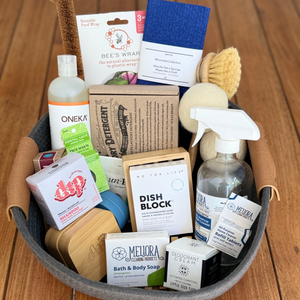A curated bath and body gift basket arranged in a gray fabric basket on a wooden deck. The basket contains a variety of personal care products including Oneka shower wrap, Dish Block, Melura bath & body soap, and other natural cleaning items.