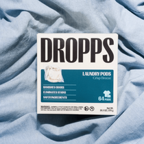 A package of DROPPS Laundry Pods in Crisp Breeze scent, placed on a rumpled light blue fabric background.