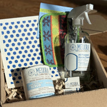 Eco-friendly cleaning kit including a glass bottle of all-purpose cleaner with it refill tablets, a scrub powder, a swedish cloth with blue dots and a scrubbing sponge, all in a box on a wooden table