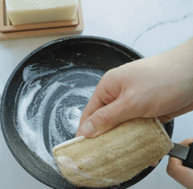 A hand holding a eco-sponge over a frying pan with soap suds.