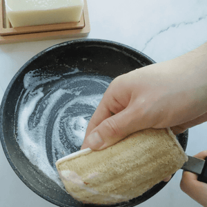 A hand holding a eco-sponge over a frying pan with soap suds.