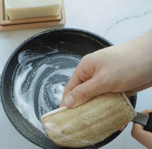 A hand holding a eco-sponge over a frying pan with soap suds.