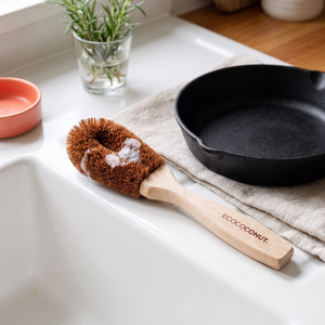 A wooden-handled scrubbing brush with coconut bristles and white soap lather rests on a white sink beside a black cast-iron skillet on a beige cloth. The handle is branded ECOCOCONUT.