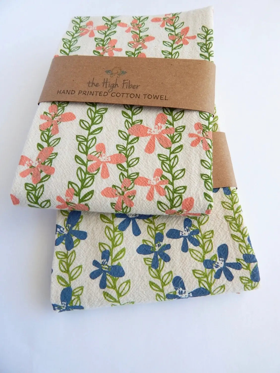 Two folded cotton towels with floral prints, wrapped together with a brown kraft paper band that reads 'the High Fiber HAND PRINTED COTTON TOWEL'. The top towel features coral-pink flowers and green vines on an off-white background, while the bottom towel features blue flowers and green vines on a similar off-white background.