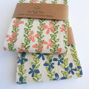 Two folded cotton towels with floral prints, wrapped together with a brown kraft paper band that reads 'the High Fiber HAND PRINTED COTTON TOWEL'. The top towel features coral-pink flowers and green vines on an off-white background, while the bottom towel features blue flowers and green vines on a similar off-white background.