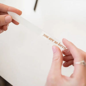 Hand holding a glass nail file with 'you are so loved' engraved on it