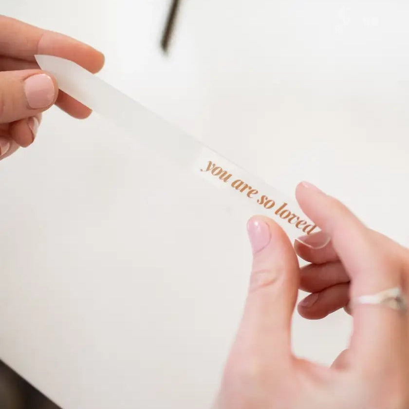 Hand holding a glass nail file with 'you are so loved' engraved on it