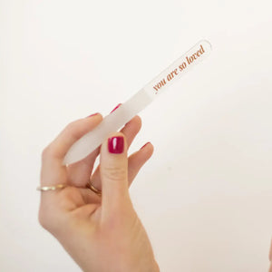 Hand holding a glass nail file with 'you are so loved' text engraved on it