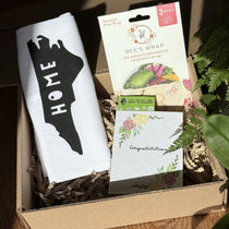 Box containing a towel with 'HOME' print, a beeswax wrap package, and a plantable seed card with the word congratulations on it.