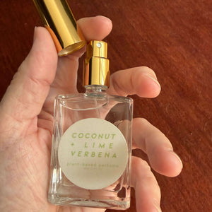 A hand holding a clear glass perfume bottle with a gold spray top and a gold cap, featuring a round cream label that reads 'COCONUT + LIME + VERBENA' and 'plant-based perfume'.