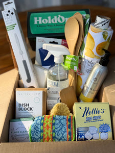 A cardboard gift box filled with assorted kitchen and cleaning products, including a spray bottle, dish block, dish cubes, a wooden spoon, a box of nut butter, and other household items.