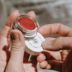 A close-up of hands holding a small round cosmetic pot with a clear lid and a white base, showing a warm coral-red cream product inside. The person is applying or swabbing the product with a small applicator on the white lid.