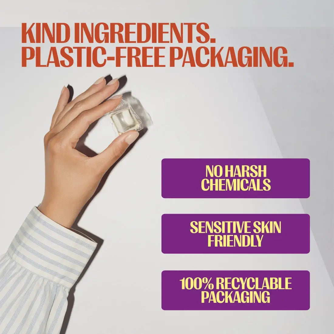 A hand wearing a light blue and white striped shirt holds a small clear plastic packaging cube against a light gray background. Large orange text at the top reads 'KIND INGREDIENTS.