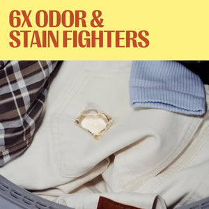 A close-up of folded clothing in a laundry basket with a clear square patch showing a detergent crystal, under a yellow banner with red text reading '6X ODOR & STAIN FIGHTERS'.