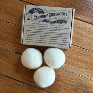 Three white wool dryer balls next to a kraft paper box of 'Laundry Detergent' by 'The Laundry Detergent Company' on a wooden floor.