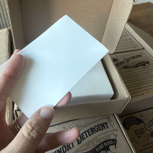 A hand holding a laundry detergent sheet in front of a cardboard box filled with laundry sheets, with branded laundry detergent boxes visible in the background.