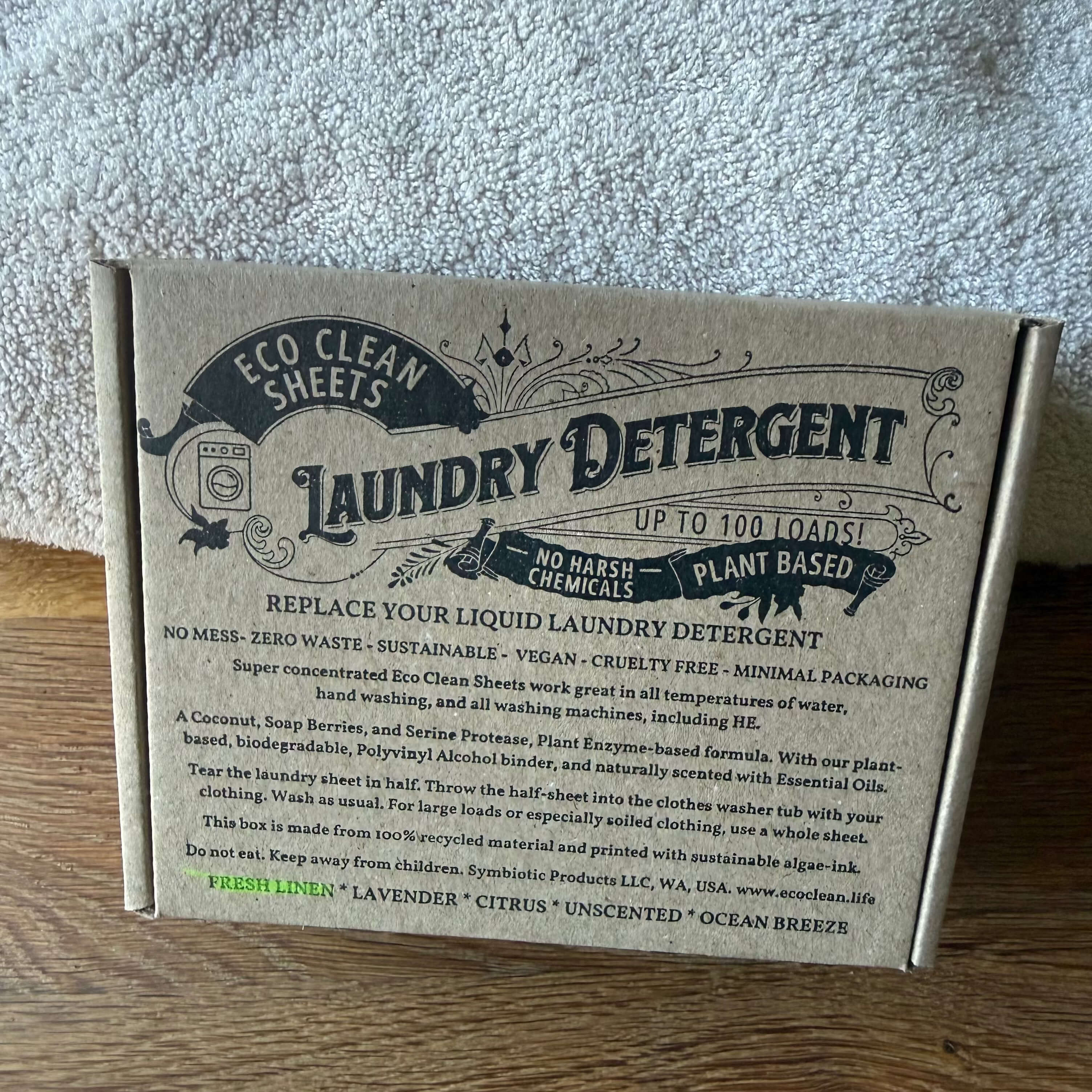 A cardboard box of Eco Clean Sheets Laundry Detergent, showing the front packaging with product details, set on a wooden surface against a gray textured background.