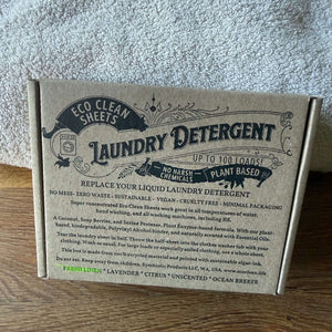 A cardboard box of Eco Clean Sheets Laundry Detergent, showing the front packaging with product details, set on a wooden surface against a gray textured background.