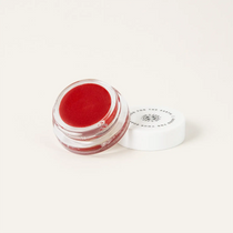 A small clear cosmetic jar filled with bright red cream or balm, shown open with its white lid placed beside it on a plain light background.
