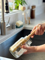 A person holding a glass jar brush with a light wood handle in a kitchen sink, with a stainless steel faucet and countertop in the background.