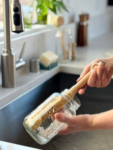 A person holding a glass jar brush with a light wood handle in a kitchen sink, with a stainless steel faucet and countertop in the background.