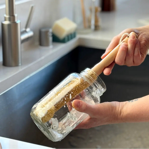 A person holding a glass jar brush with a light wood handle in a kitchen sink, with a stainless steel faucet and countertop in the background.