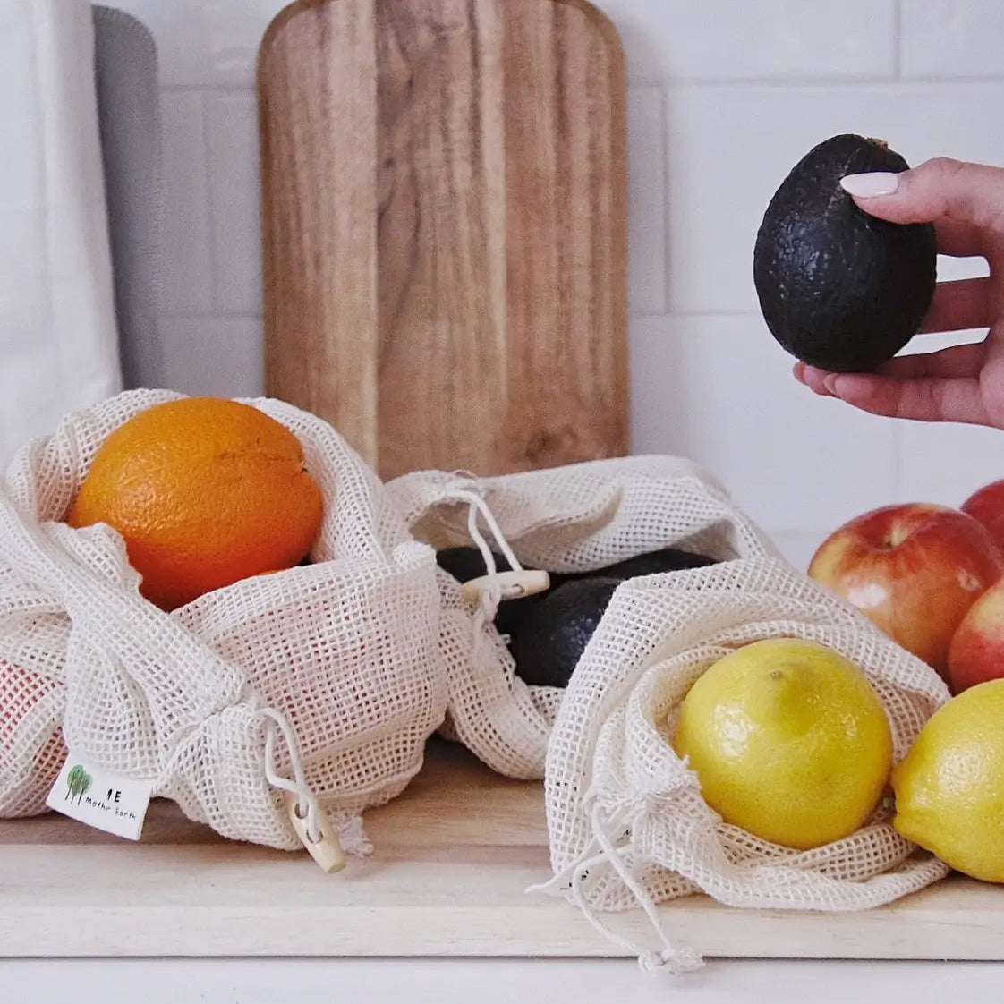 A hand holds a dark avocado above reusable beige mesh drawstring bags containing an orange, more avocados, a yellow pear, and lemons, with red apples in the background and a wooden cutting board against a white tiled wall.