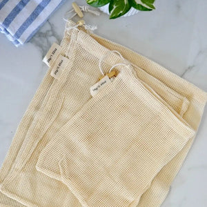 Set of beige mesh bags on a marble surface with a plant and towel in the background.