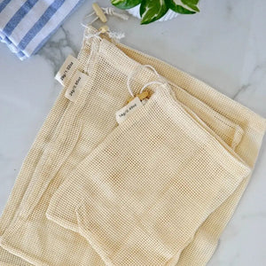 Set of beige mesh bags on a marble surface with a plant and towel in the background.
